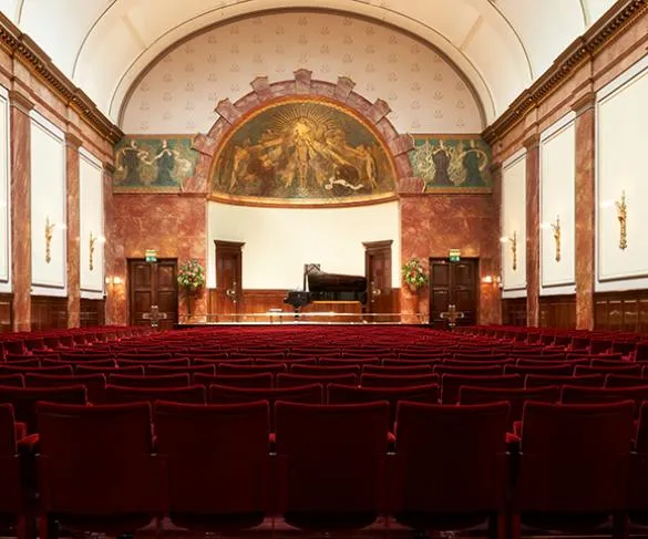 A grand concert hall with a stage featuring a piano, surrounded by red velvet seats and ornate wall decorations.