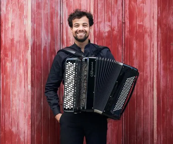 Man in a black shirt smiling and holding an accordion in front of a red wooden wall.