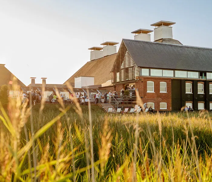 A group of people socializes on a patio at a modern building surrounded by tall grass under a clear sky.