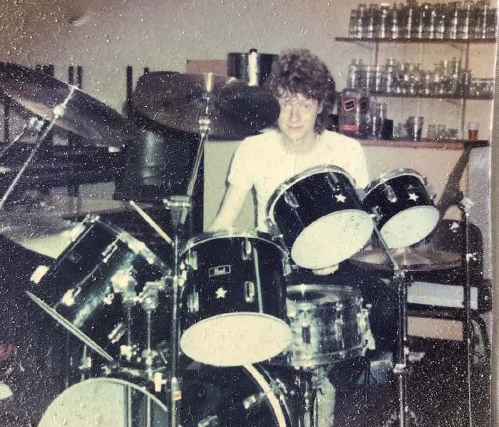 Person playing a drum set in a room with shelves holding various jars and containers.