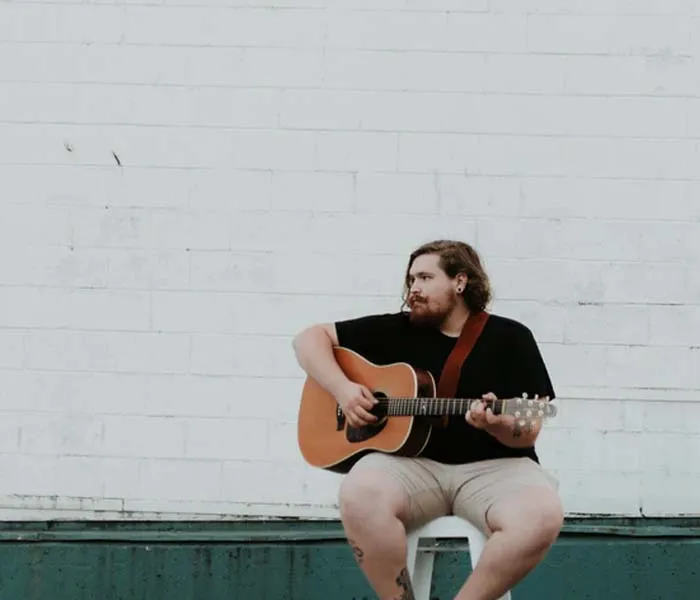 Person with a beard sitting on a stool, playing an acoustic guitar against a white brick wall background.