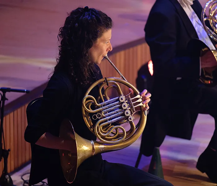 Woman playing a French horn in an orchestra, seated on a wooden stage, wearing a black outfit.