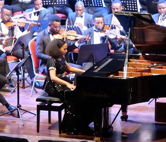 A woman in a black dress plays a grand piano on stage, accompanied by an orchestra of musicians in suits.