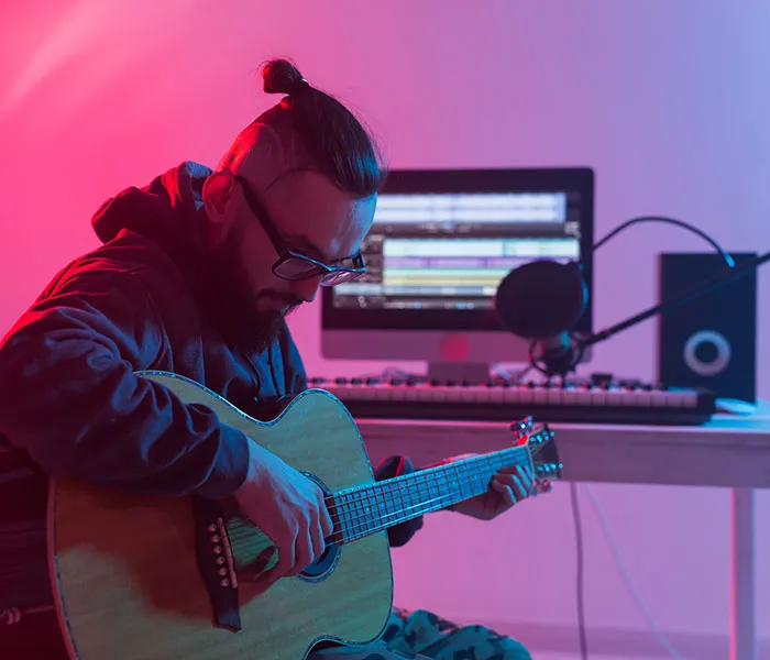 Person with glasses and tied hair plays an acoustic guitar in a room with a computer and audio equipment.