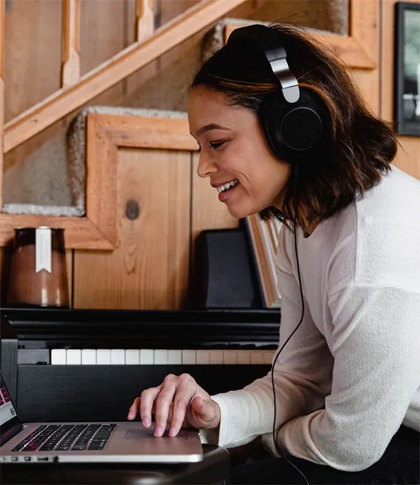 A person wearing headphones smiles while using a laptop in front of a piano.