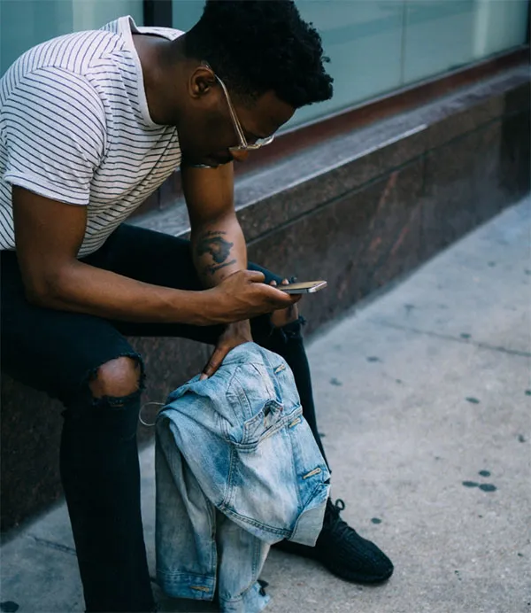 Person sitting on a sidewalk, looking at their phone, holding a denim jacket, wearing a striped shirt and black pants.