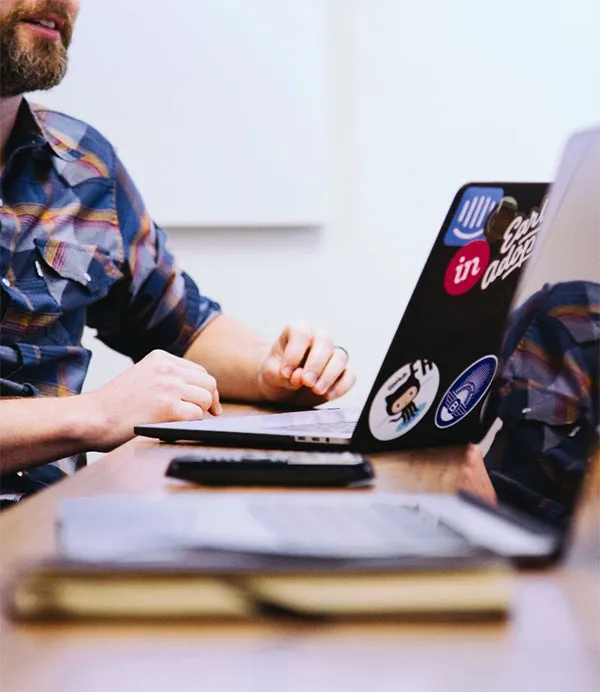 Man seated at a table with a sticker-covered laptop, a closed notebook, and a smartphone nearby.