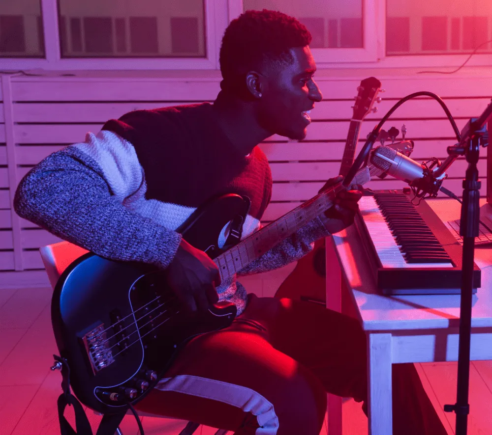 Person playing electric guitar and singing into a microphone at a desk with a keyboard, under pink and purple lighting.