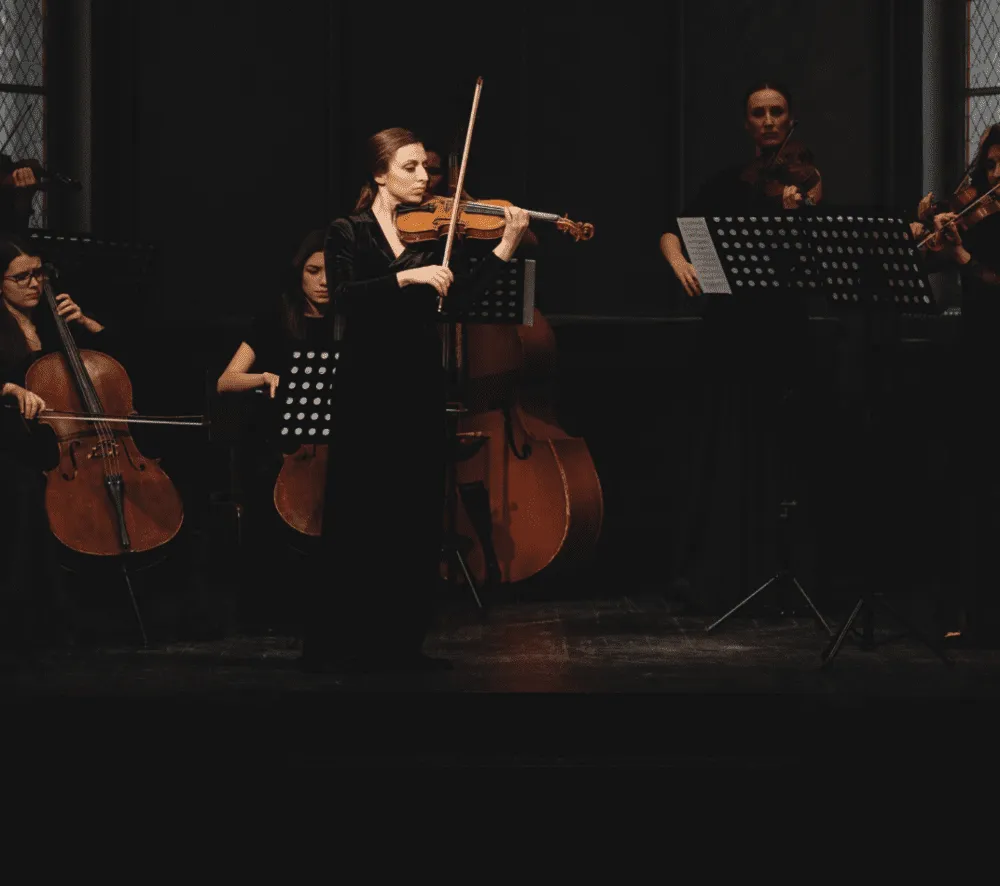 A violinist in a black dress performs on stage with an orchestra, featuring cellists and sheet music stands.