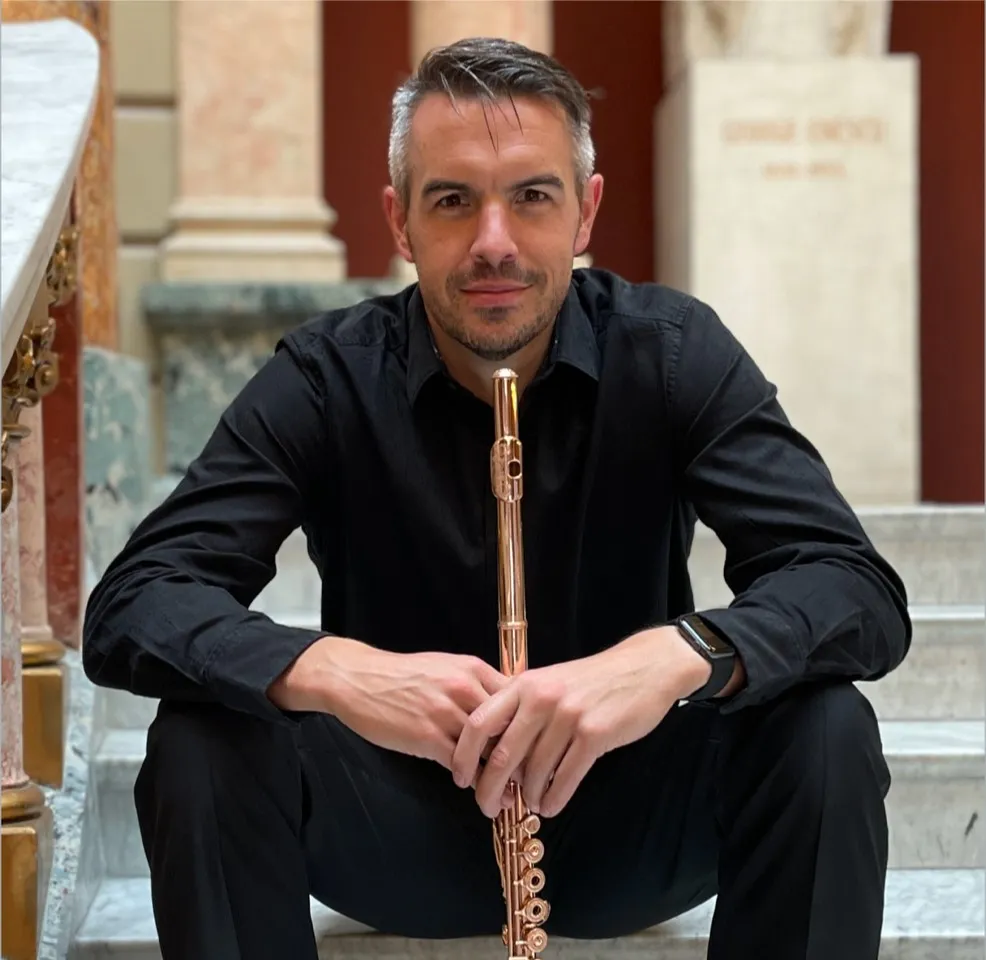 Man seated on stairs holding a flute, wearing a black shirt. Marble columns and a large statue in the background.