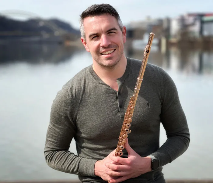 Man in a gray shirt holds a flute, standing in front of a calm lake with a bridge and buildings in the background.