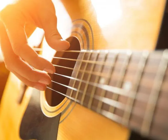 Close-up of a person’s hand strumming the strings of an acoustic guitar, with sunlight casting a warm glow on the instrument.