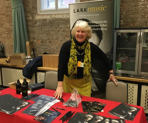 A woman stands behind a table with promotional materials for Lark Music, smiling. The setup is in front of a brick wall.