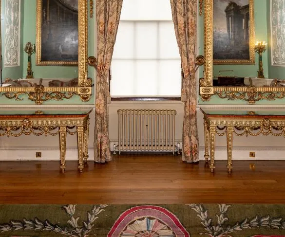 A room with ornate gold-framed mirrors, floral curtains, two decorative tables, and a radiator centered below a large window.