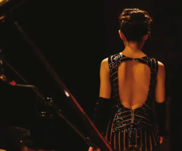 A woman in a backless, sequined dress stands on stage facing away, with dim lighting highlighting her elegant attire.