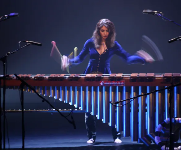 A person plays a large marimba on stage using multiple mallets, with microphones surrounding the instrument.