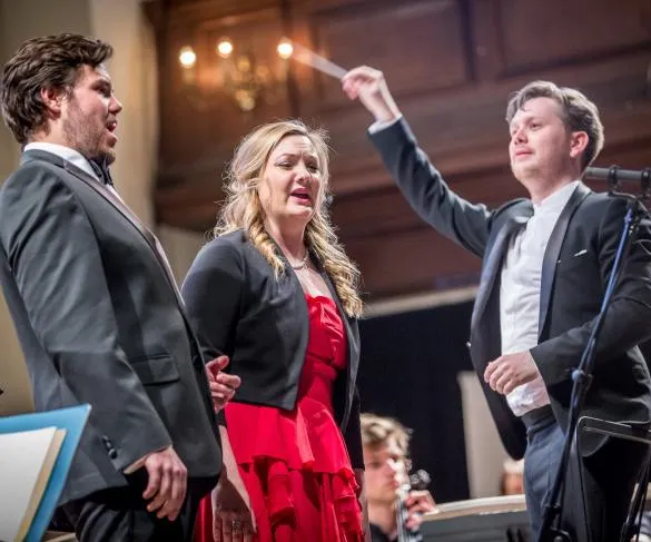 Two singers perform on stage while a conductor leads the orchestra in a formal concert setting.