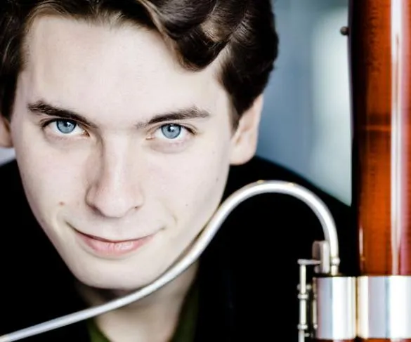 A person with blue eyes and brown hair is looking at the camera while holding a bassoon.