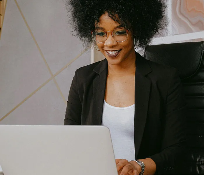 A person wearing glasses and a black blazer is smiling while working on a laptop at a desk.