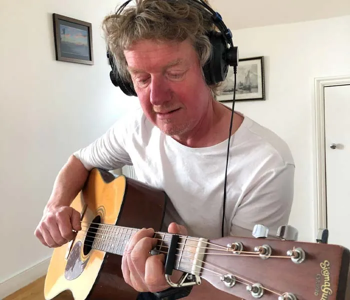 A man with headphones is playing an acoustic guitar in a room with framed pictures on the wall.