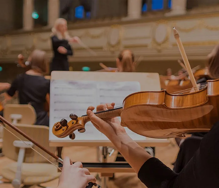 Close-up of a violinist playing in an orchestra, with the conductor and sheet music stand visible in the background.