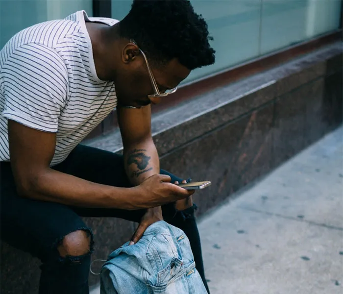 A man in a striped shirt and ripped jeans sits on a curb, looking at his phone with a backpack placed on the ground beside him.