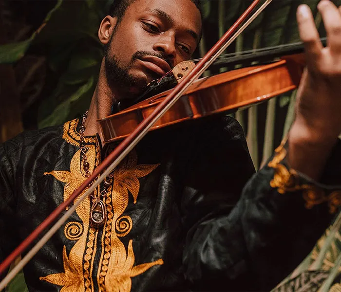 Person wearing a decorative shirt playing a violin with focus and concentration, surrounded by green foliage.