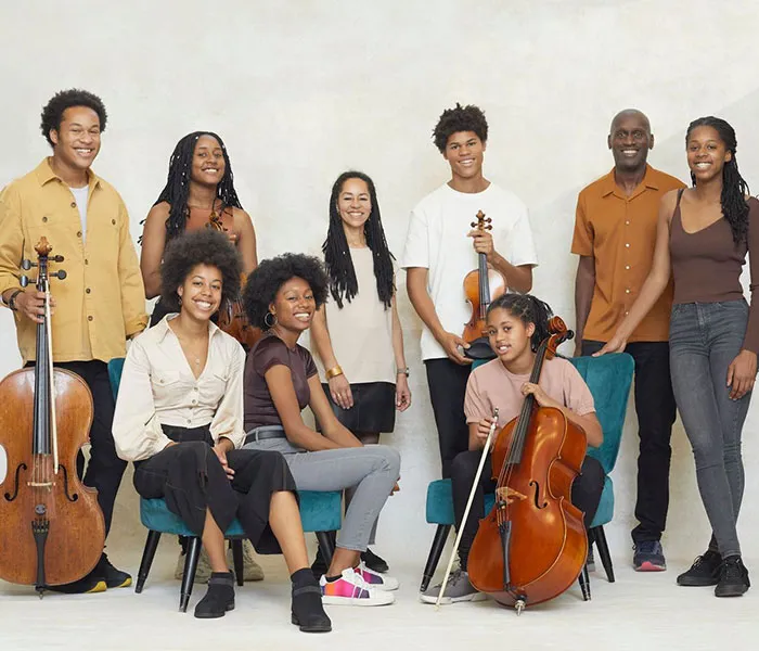 A group of nine people, including adults and children, posed together with musical instruments such as cellos and a violin.