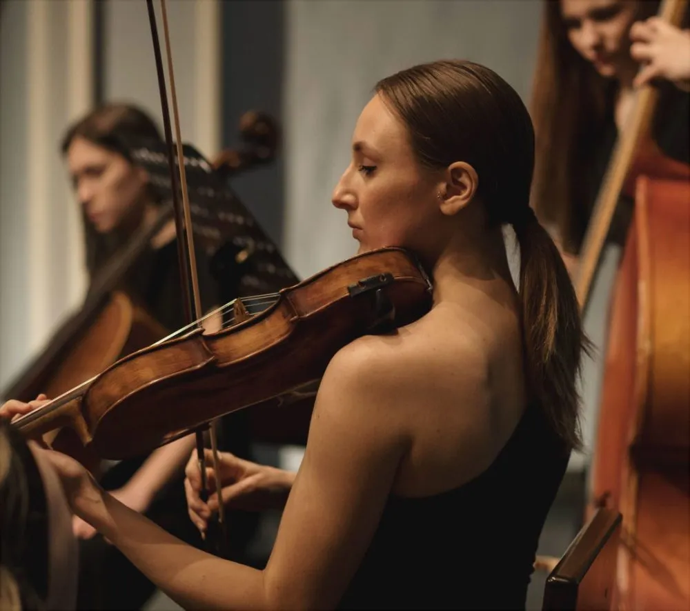 A focused violinist plays her instrument during a performance, with other musicians visible in the background.