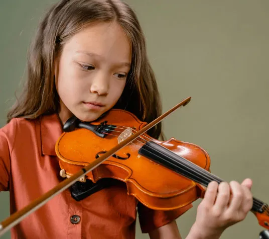 A young girl with long hair plays the violin while looking down, dressed in an orange shirt, against a green background.