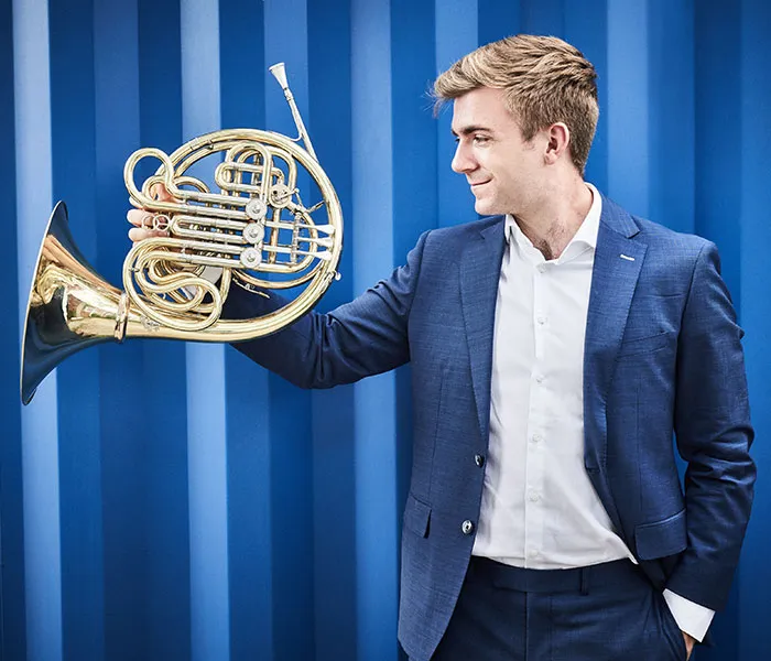 Man in a blue suit holding a French horn against a blue striped background, looking at the instrument.