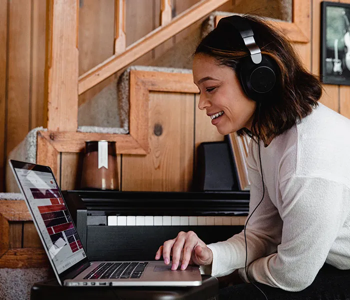 Person wearing headphones smiles while using a laptop, sitting near a staircase and a piano in a wood-paneled room.