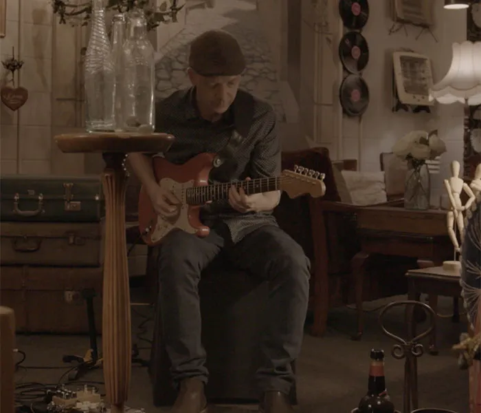 A man wearing a hat plays an electric guitar while seated in a room decorated with vintage items, including vinyl records and suitcases.