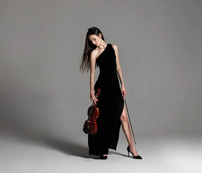 A woman in a black one-shoulder dress holds a violin and bow, standing against a plain gray background.