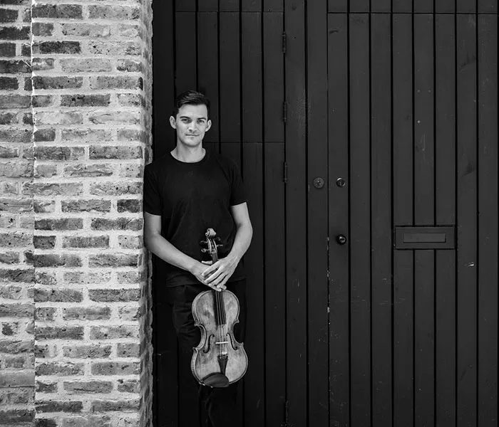 A person stands against a brick wall holding a violin in front of a wooden door.