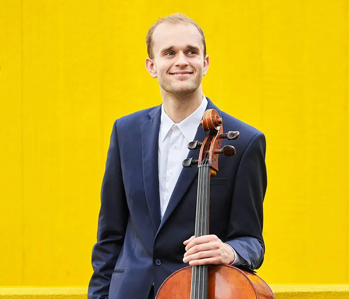 A person in a blue suit holds a cello upright with a yellow background.