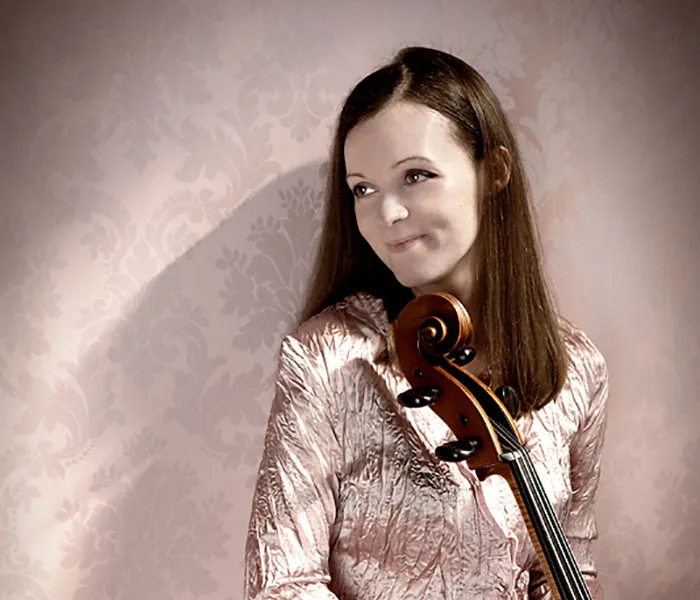 A woman with long brown hair in a pink metallic top holds a cello, standing against a light pink patterned background.