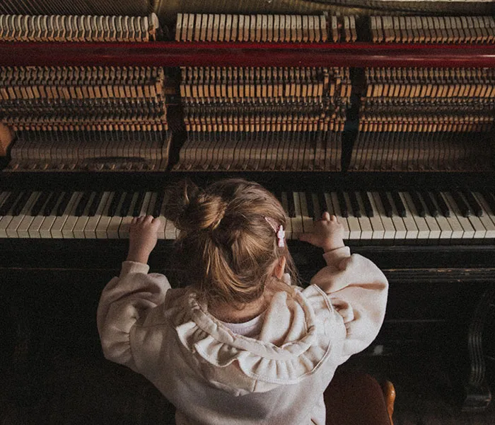 A young child with a hair clip stands playing a piano, viewed from behind, showing the inner mechanics of the instrument.