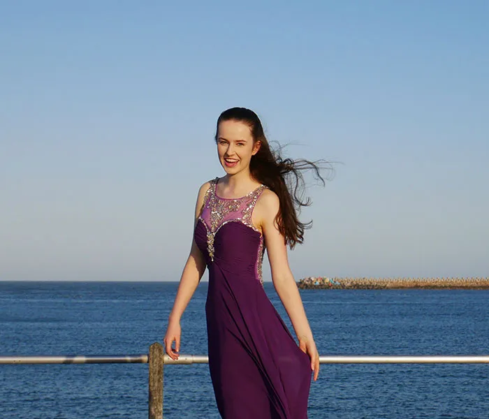 A woman in a purple dress stands by the seaside, smiling, with the ocean and clear blue sky in the background.