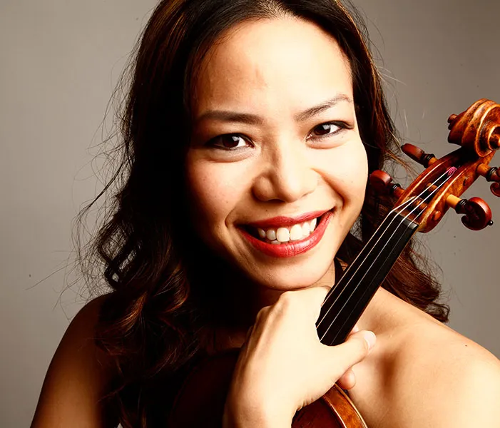 A person with shoulder-length dark hair smiles and holds a violin close to their face against a plain background.