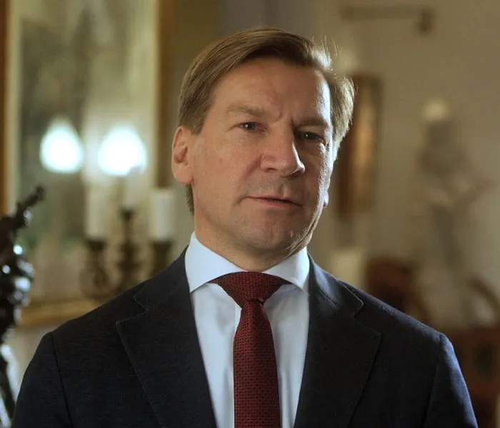 A man in a suit and red tie stands indoors, looking forward. The background shows a blurred interior with decor.