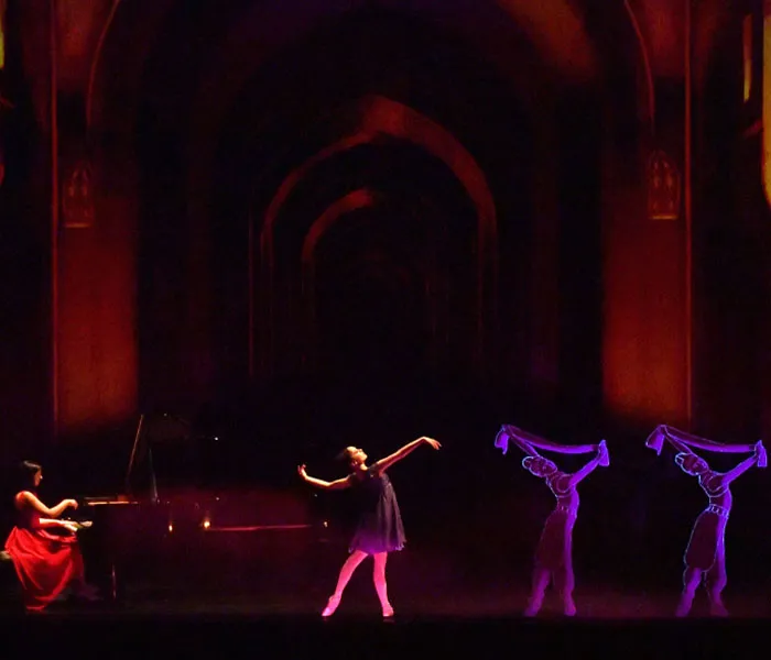 A pianist in red plays on stage while a ballerina dances in the center; two dancers with glowing scarves move in the background.