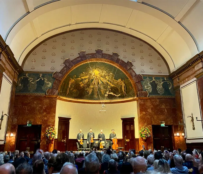 Four musicians perform on a stage under an ornate arch in a packed concert hall.