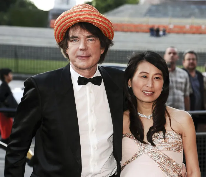 A man in a black tuxedo and orange striped hat poses with a woman in a light-colored dress at an outdoor event.