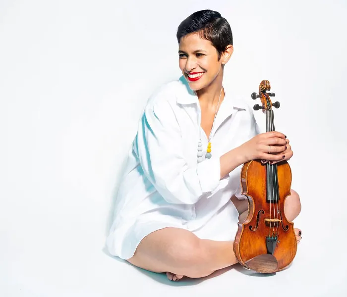 A person with short hair sits on the floor, smiling and holding a violin, wearing a white shirt and a colorful necklace.