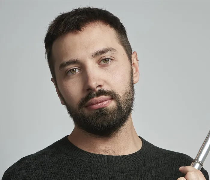 A man with short dark hair and a beard, wearing a dark pullover, looks directly at the camera against a plain gray background.