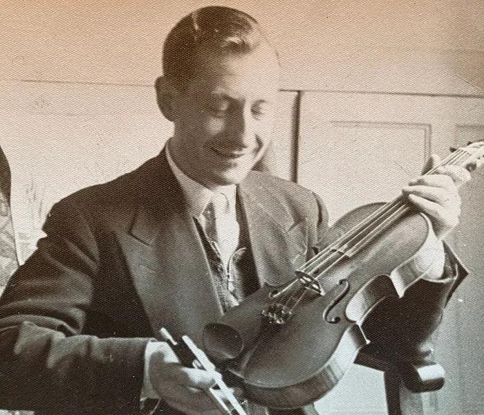 A man in a suit smiling while holding a violin and bow, seemingly preparing to play the instrument.