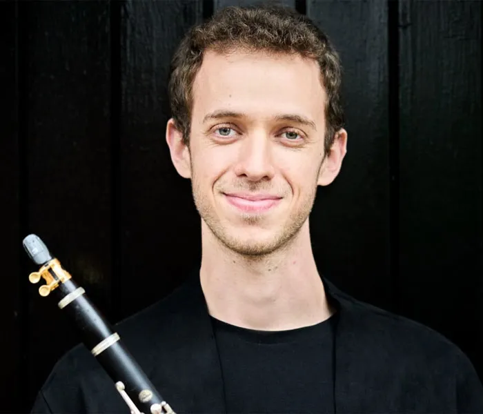 A man stands against a dark background, holding a clarinet and smiling at the camera, dressed in a black shirt.