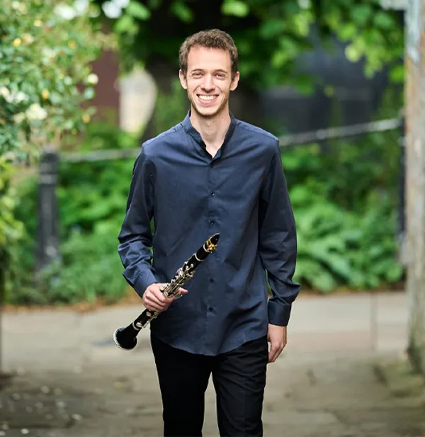 A person wearing a dark shirt and pants smiles while walking outdoors in a green setting, holding a clarinet with their right hand.