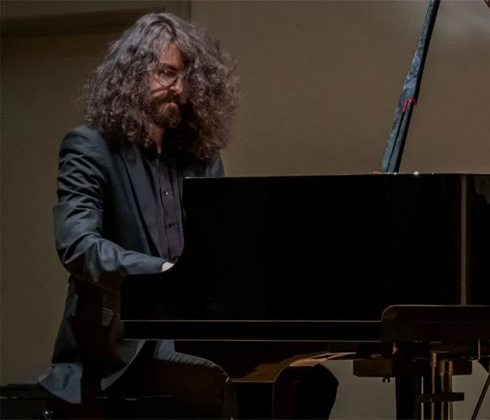 A person with long, curly hair and glasses is playing a grand piano while seated, dressed in a dark suit.